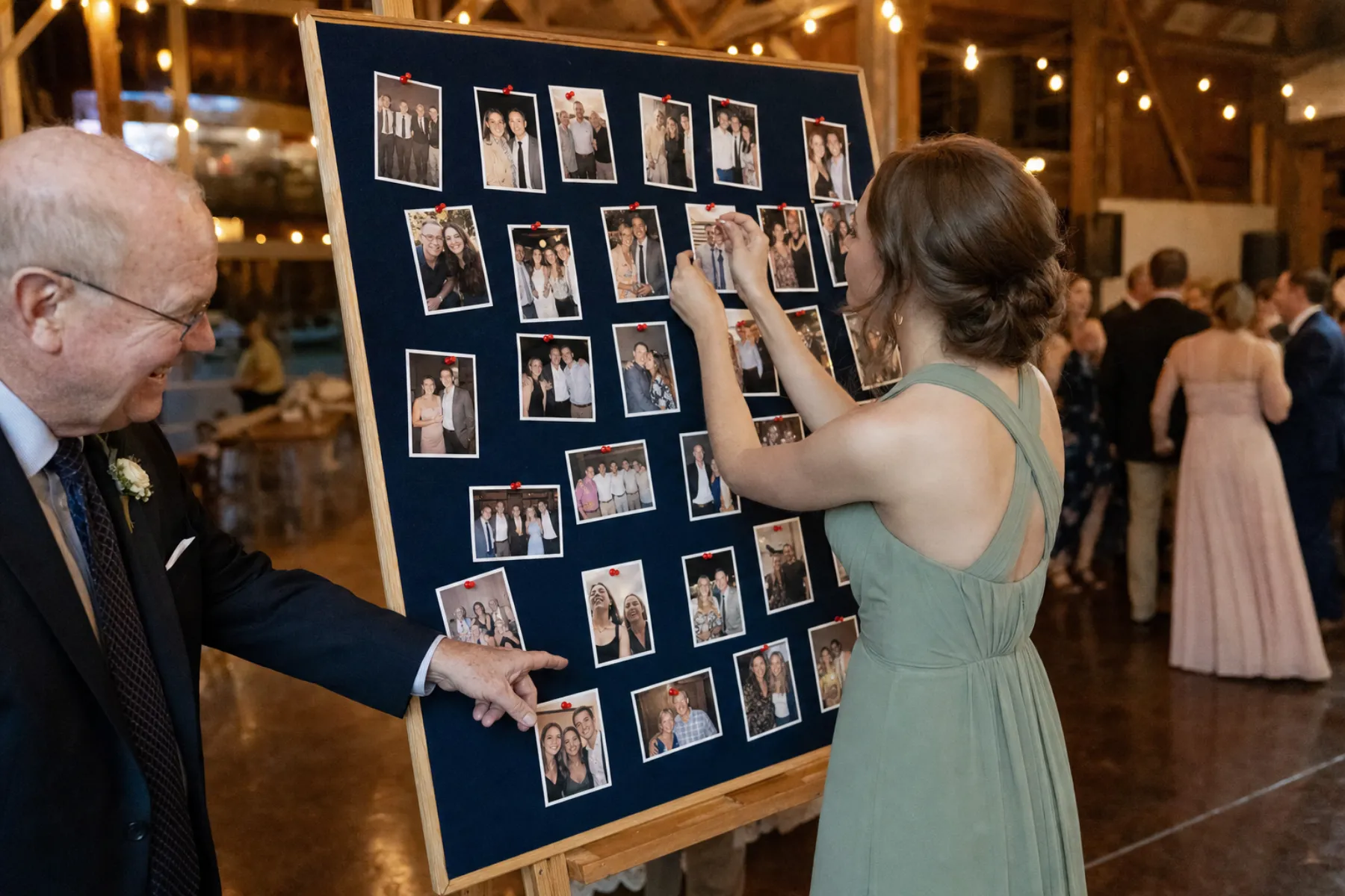 A bridesmaid pinning a print to a navy board on an easel at a barn wedding while a guest points at a photo